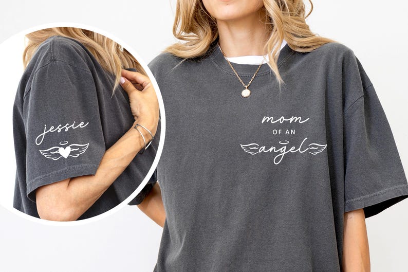 May include: Dark gray t-shirt with the words "mom of an angel" and a heart with wings graphic. The sleeve has the name "Jessie" and a similar heart and wing design. The shirt has a relaxed fit.
