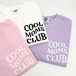 May include: Three t-shirts with the text "Cool Moms Club" printed on them. One shirt is white with black text, one is pink with white text, and one is lavender with white text.