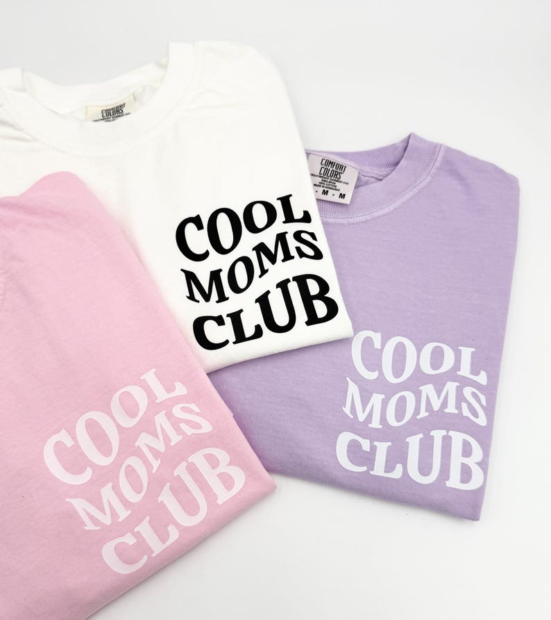 May include: Three t-shirts with the text "Cool Moms Club" printed on them. One shirt is white with black text, one is pink with white text, and one is lavender with white text.