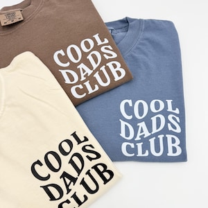 Pode incluir: Tr&ecirc;s camisetas com o texto "Cool Dads Club" em uma fonte estilizada. As camisetas s&atilde;o de cor marrom, azul claro e creme.