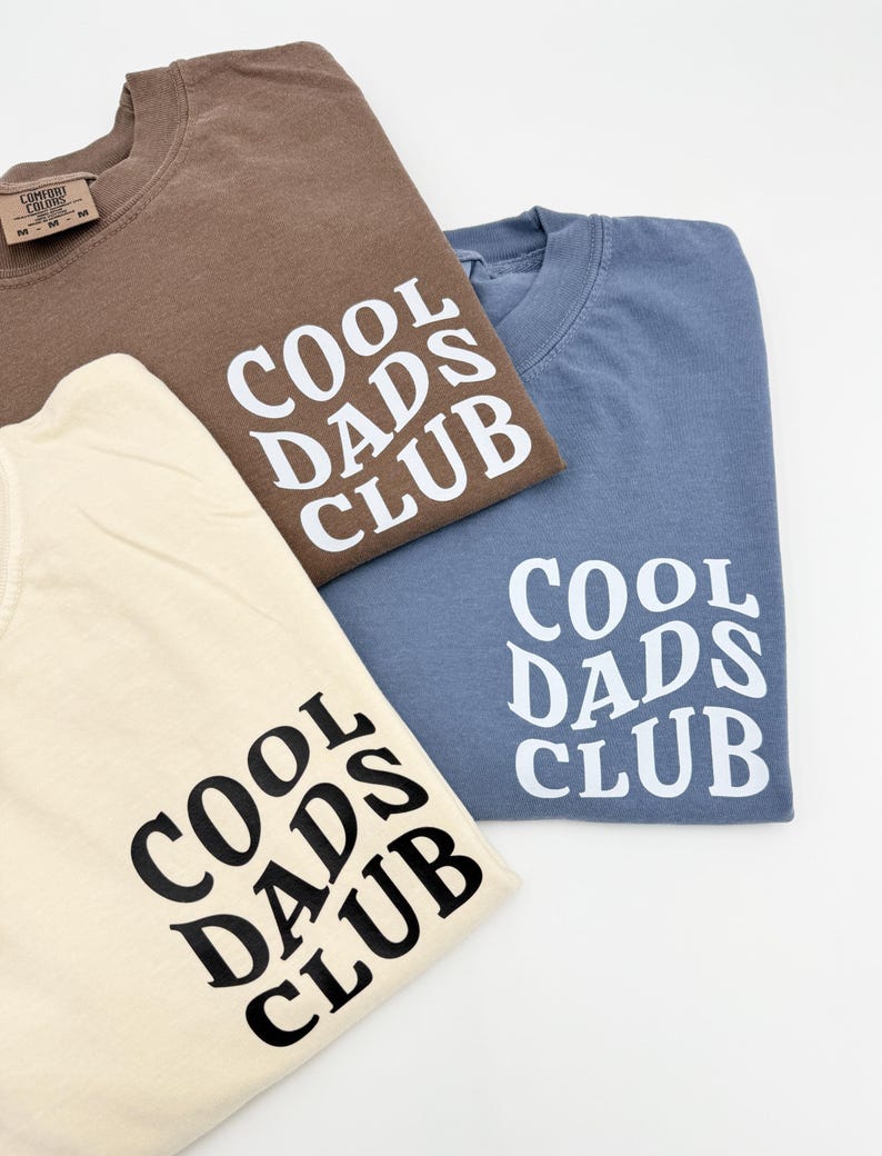 Pode incluir: Tr&ecirc;s camisetas com o texto "Cool Dads Club" em uma fonte estilizada. As camisetas s&atilde;o de cor marrom, azul claro e creme.