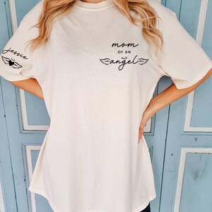 May include: Cream-colored t-shirt with the text "mom of an angel" and the name "Jessie" printed on it. The shirt features a black heart and wing design. The shirt is a casual, oversized fit.