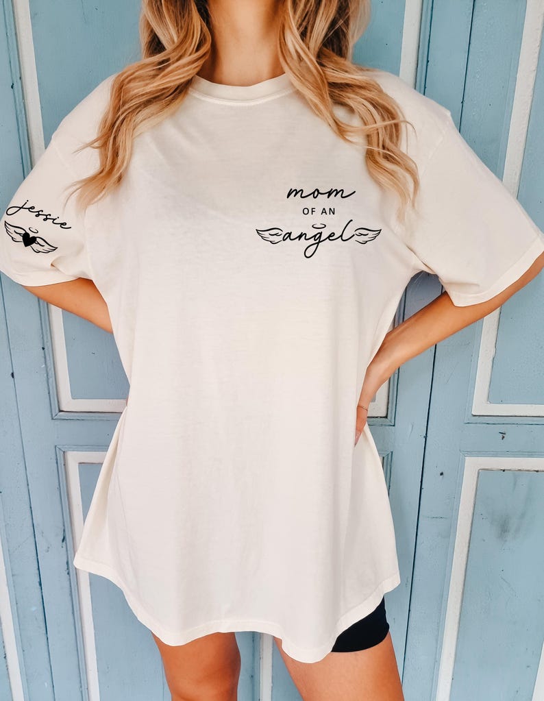 May include: Cream-colored t-shirt with the text "mom of an angel" and the name "Jessie" printed on it. The shirt features a black heart and wing design. The shirt is a casual, oversized fit.