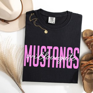 Puede incluir: Camiseta negra con la palabra "MUSTANGS" en letras rosas y "willowgate" en cursiva. Un collar dorado, un sombrero beige y sandalias marrones tambi&eacute;n est&aacute;n en la imagen.