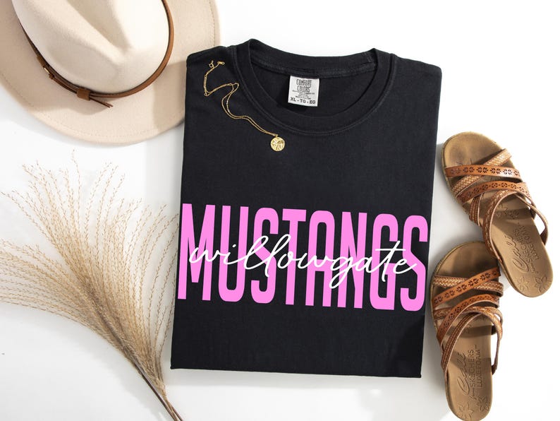 Puede incluir: Camiseta negra con la palabra "MUSTANGS" en letras rosas y "willowgate" en cursiva. Un collar dorado, un sombrero beige y sandalias marrones tambi&eacute;n est&aacute;n en la imagen.