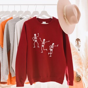 May include: A red sweatshirt featuring a white graphic of three skeletons dancing. The sweatshirt is hanging on a hanger with other clothing.