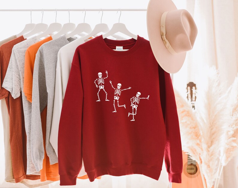 May include: A red sweatshirt featuring a white graphic of three skeletons dancing. The sweatshirt is hanging on a hanger with other clothing.
