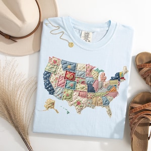 May include: Light blue t-shirt with an embroidered patchwork map of the United States. The shirt is next to a beige hat with a brown band, a gold necklace, and brown leather sandals. A bundle of dried ornamental grass is also present.