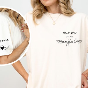 May include: Cream-colored t-shirt with the text "mom of an angel" in black script and a pair of angel wings. The left sleeve has the name "Jessie" and a heart with wings graphic. The shirt is a casual style.