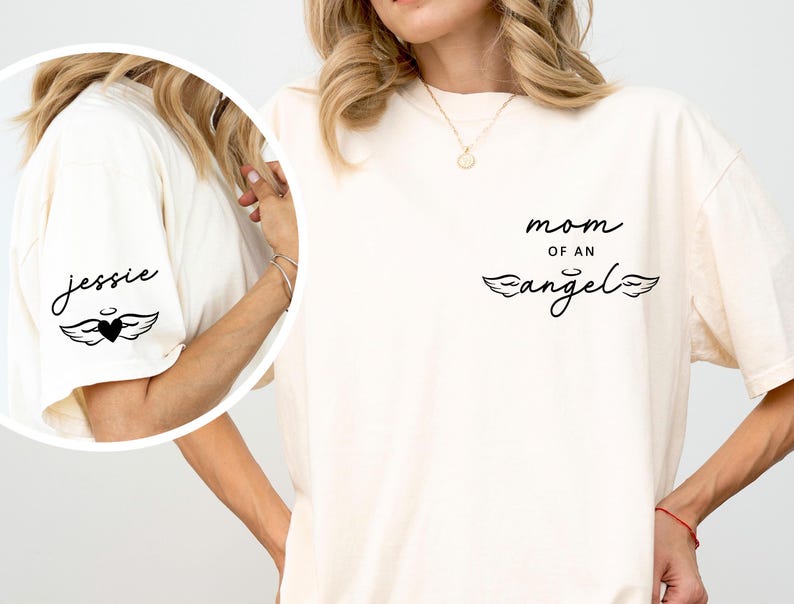 May include: Cream-colored t-shirt with the text "mom of an angel" in black script and a pair of angel wings. The left sleeve has the name "Jessie" and a heart with wings graphic. The shirt is a casual style.
