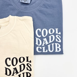 Pode incluir: Duas camisetas com o texto "Cool Dads Club" impresso na manga. Uma camiseta &eacute; azul claro e a outra &eacute; creme.