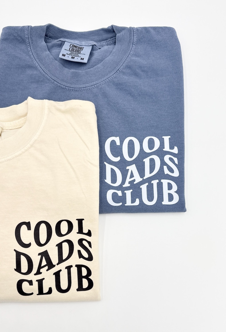 Pode incluir: Duas camisetas com o texto "Cool Dads Club" impresso na manga. Uma camiseta &eacute; azul claro e a outra &eacute; creme.