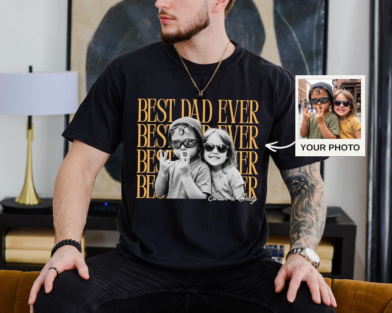 A man wearing a black t-shirt with a graphic design featuring two people and text, sitting on a couch.
