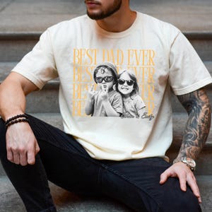 A man wearing a white t-shirt with a graphic design featuring two people, one of whom is wearing sunglasses.