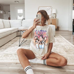 May include: A woman wearing a light beige t-shirt with a graphic print of a group of women and the text "CATIE" in pink and blue letters. The graphic print has a sunset background with pink, orange, and yellow colors. The woman is sitting on a light brown wooden floor with her legs crossed. She is wearing white socks with black stripes.
