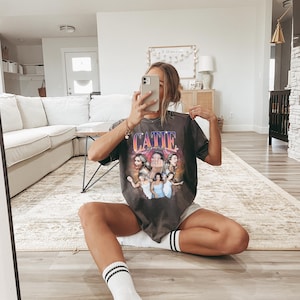 May include: A woman wearing a gray t-shirt with a graphic print of a group of women and the text "CATIE" on the front. She is sitting on a rug with her legs crossed and wearing white socks with black stripes.
