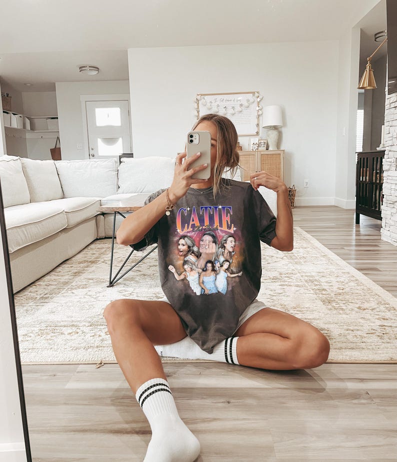 May include: A woman wearing a gray t-shirt with a graphic print of a group of women and the text "CATIE" on the front. She is sitting on a rug with her legs crossed and wearing white socks with black stripes.