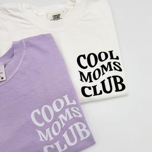 May include: Two t-shirts with the text "Cool Moms Club" printed on them. One shirt is white with black text and the other is lavender with white text.