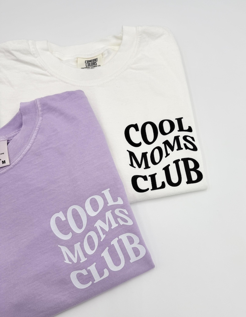 May include: Two t-shirts with the text "Cool Moms Club" printed on them. One shirt is white with black text and the other is lavender with white text.