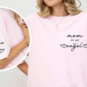 May include: A light pink t-shirt with the words "mom of an angel" in black script, along with a heart and wing design. The sleeve has the name "jessie" in script with a heart and wing design.