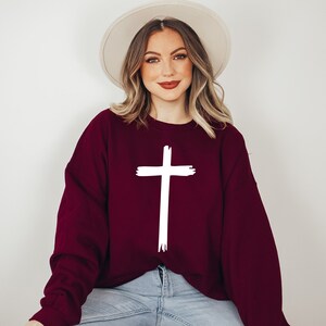 May include: A maroon sweatshirt with a white cross graphic. The sweatshirt has long sleeves and a relaxed fit.
