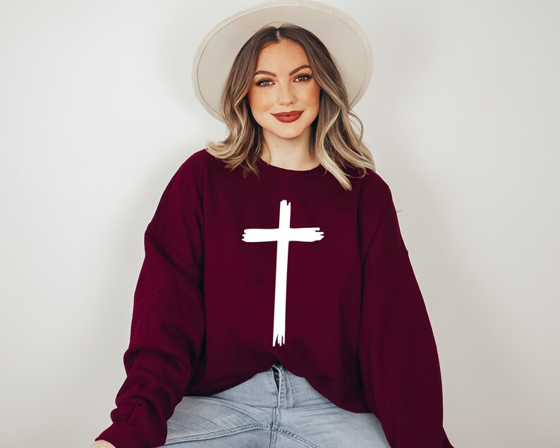 May include: A maroon sweatshirt with a white cross graphic. The sweatshirt has long sleeves and a relaxed fit.