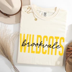 Puede incluir: Camiseta color crema con las palabras "WILDCATS" en letras amarillas y "brookvale" en escritura negra. Un collar dorado, un sombrero beige y sandalias marrones tambi&eacute;n est&aacute;n en la imagen.