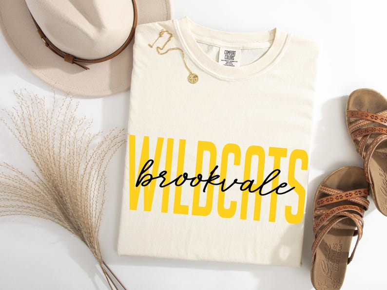 Puede incluir: Camiseta color crema con las palabras "WILDCATS" en letras amarillas y "brookvale" en escritura negra. Un collar dorado, un sombrero beige y sandalias marrones tambi&eacute;n est&aacute;n en la imagen.