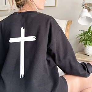 May include: Black sweatshirt with a white cross graphic on the back. The cross is drawn with a brushstroke effect.