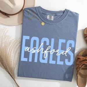 Puede incluir: Una camiseta azul con la palabra "EAGLES" en letras may&uacute;sculas azul claro y "ashford" en escritura blanca. La camiseta est&aacute; junto a un par de sandalias marrones, un sombrero beige y un collar dorado.