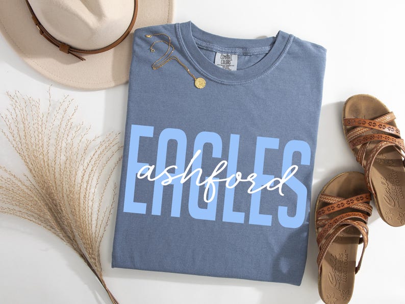 Puede incluir: Una camiseta azul con la palabra "EAGLES" en letras may&uacute;sculas azul claro y "ashford" en escritura blanca. La camiseta est&aacute; junto a un par de sandalias marrones, un sombrero beige y un collar dorado.