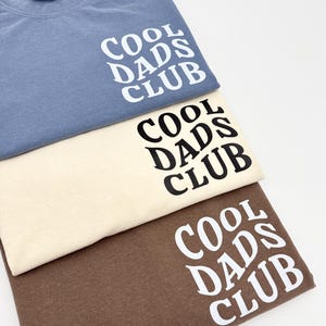 Pode incluir: Tr&ecirc;s t-shirts em cores diferentes: azul, bege e marrom. Cada camiseta tem o texto "COOL DADS CLUB" impresso na frente em uma fonte preta e em negrito.