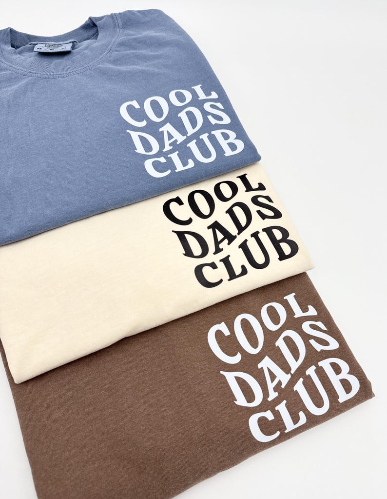 Pode incluir: Tr&ecirc;s t-shirts em cores diferentes: azul, bege e marrom. Cada camiseta tem o texto "COOL DADS CLUB" impresso na frente em uma fonte preta e em negrito.