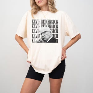 A woman wearing a white t-shirt with a black and white graphic of a man's face and the text "KEVIN REDDINGTON" repeated around the image.