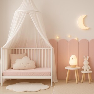 Pode incluir: Um quarto de crian&ccedil;a com um ber&ccedil;o branco, dossel e almofada em forma de nuvem. Um colch&atilde;o rosa e pain&eacute;is de parede complementam a decora&ccedil;&atilde;o. Uma l&acirc;mpada de coelho e um brinquedo est&atilde;o em pequenas mesas de madeira. Obras de arte e uma luz de lua crescente.