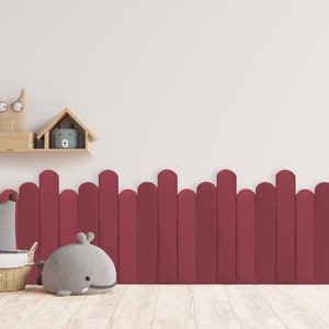 May include: A burgundy wooden fence with rounded tops, a gray whale stuffed animal, a gray pillow with brown trim, and a wicker basket with a wooden elephant toy. The background is a white wall with a wooden shelf holding a wooden house and a stuffed animal.