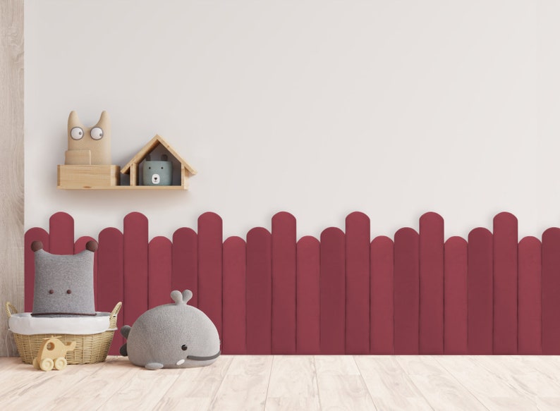 May include: A burgundy wooden fence with rounded tops, a gray whale stuffed animal, a gray pillow with brown trim, and a wicker basket with a wooden elephant toy. The background is a white wall with a wooden shelf holding a wooden house and a stuffed animal.