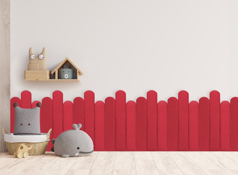 May include: A red felt fence with rounded tops, a gray whale plush toy, a gray plush toy with a brown border, and a wicker basket with a small wooden toy elephant on wheels. The toys are in front of a white wall with a wooden shelf holding a wooden house and a gray cat plush toy.