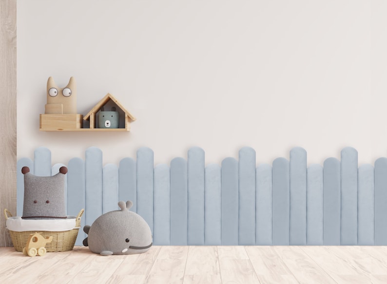 May include: A light blue wooden fence with rounded tops, a gray whale stuffed animal, a gray and brown stuffed pillow in a wicker basket, and a wooden shelf with a gray stuffed animal and a wooden house on a white wall.