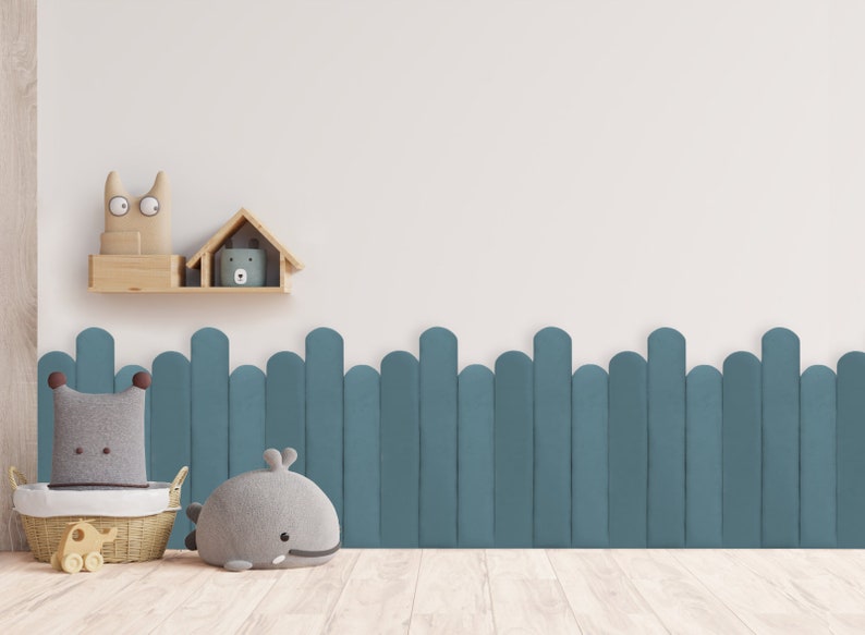 May include: A white wall with a wooden shelf holding two stuffed animal toys. The wall is decorated with a blue fence made of rounded wooden slats. A gray whale stuffed animal and a gray and brown stuffed animal are on the floor in front of the fence.