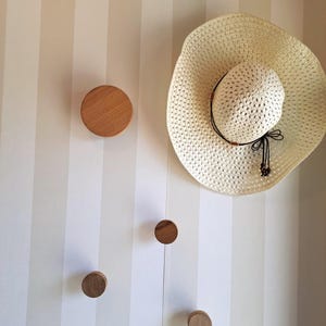 Op de afbeelding: Vier ronde houten wandhaken, gemonteerd op een wit gestreepte muur. Een witte strohoed hangt aan een van de haken.