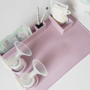 May include: A light purple silicone breast pump accessory mat featuring integrated circular holders for two pump flanges and bottles, plus two rectangular compartments for storage bags and markers. The mat has the word MOJO embossed on the surface.