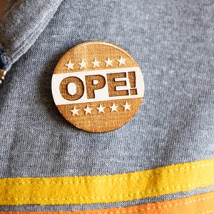May include: A wooden button with a white background and the word "OPE!" in black lettering. The button is surrounded by a ring of white stars.