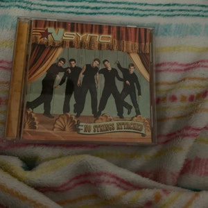 May include: A CD case with a picture of the boy band *NSYNC on the cover. The band members are dressed in black and white suits and are standing in front of a red curtain. The title of the album is "No Strings Attached".