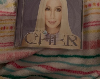 Puede incluir: Una caja de CD morada con una imagen de Cher en la parte delantera. El texto "The Very Best Of Cher" está impreso en la caja.