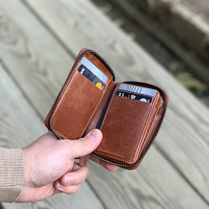 Puede incluir: Una cartera de cuero marr&oacute;n con cremallera y m&uacute;ltiples ranuras para tarjetas. La cartera est&aacute; abierta y tiene varias tarjetas dentro.