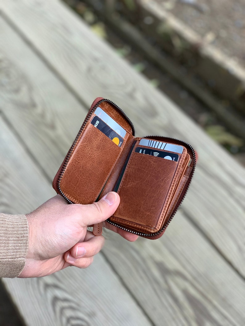 Puede incluir: Una cartera de cuero marr&oacute;n con cremallera y m&uacute;ltiples ranuras para tarjetas. La cartera est&aacute; abierta y tiene varias tarjetas dentro.