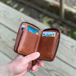 Puede incluir: Una cartera de cuero marr&oacute;n con cremallera y m&uacute;ltiples ranuras para tarjetas. La cartera est&aacute; abierta y tiene tarjetas en su interior.