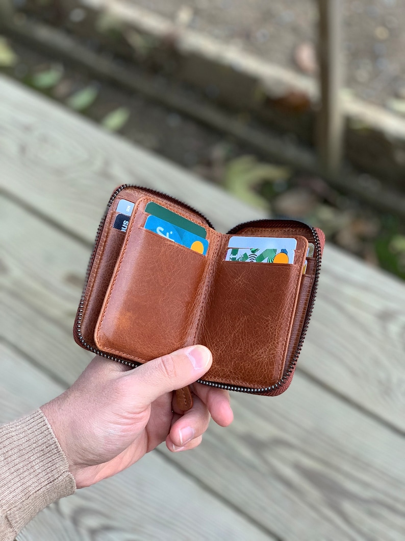 Puede incluir: Una cartera de cuero marr&oacute;n con cremallera y m&uacute;ltiples ranuras para tarjetas. La cartera est&aacute; abierta y tiene tarjetas en su interior.