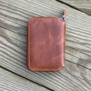 Puede incluir: Una cartera de cuero marr&oacute;n con cremallera y detalles de costura marr&oacute;n claro.